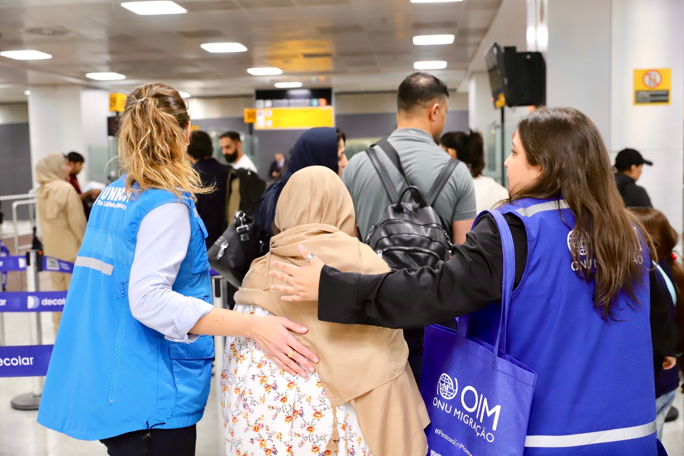 Staff from UNHCR and IOM walk through a crowded airport with their arms around a woman