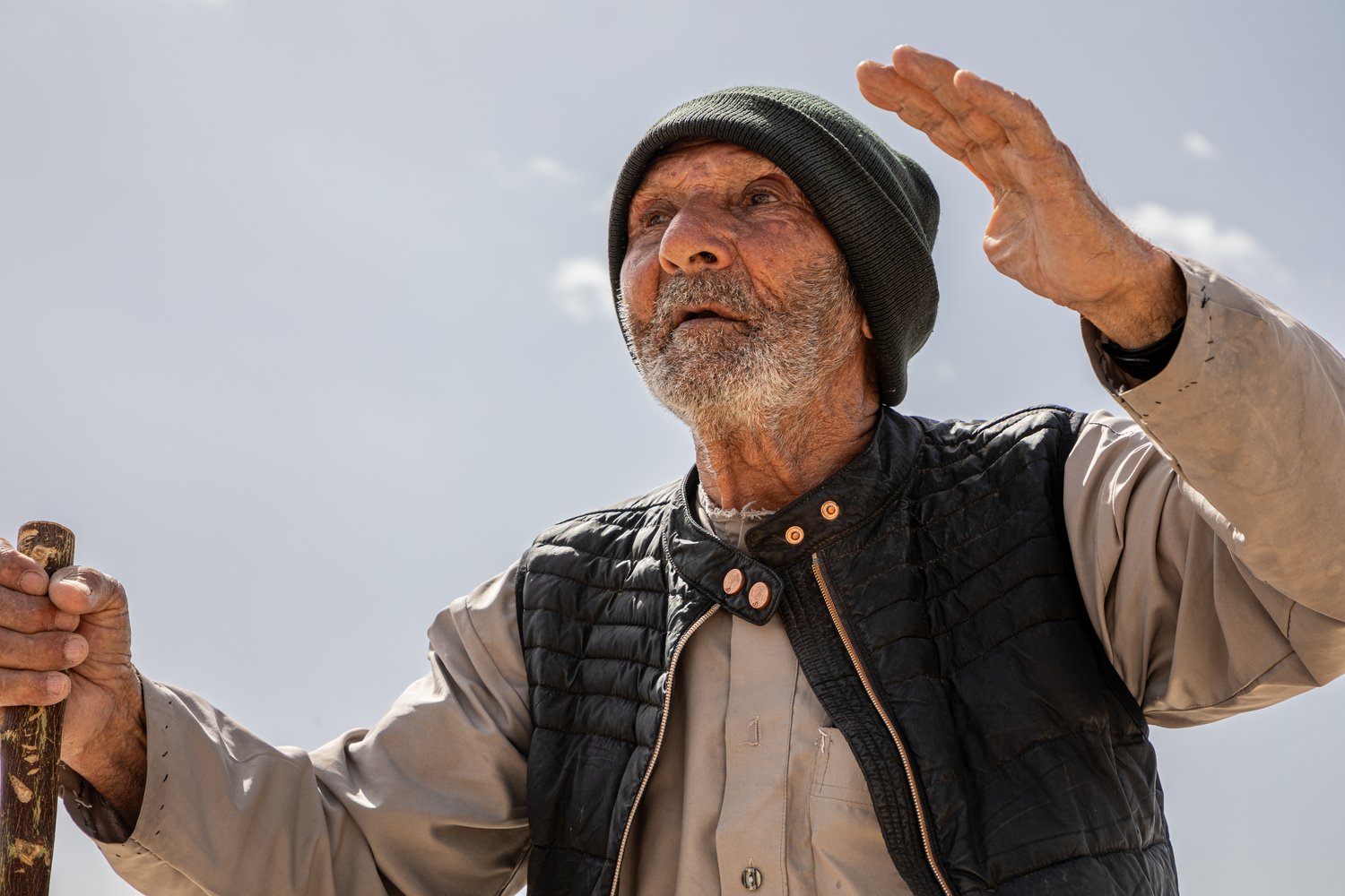 An elderly man wearing a cap and holding a walking stick stands outside beneath a clear sky with his hands raised