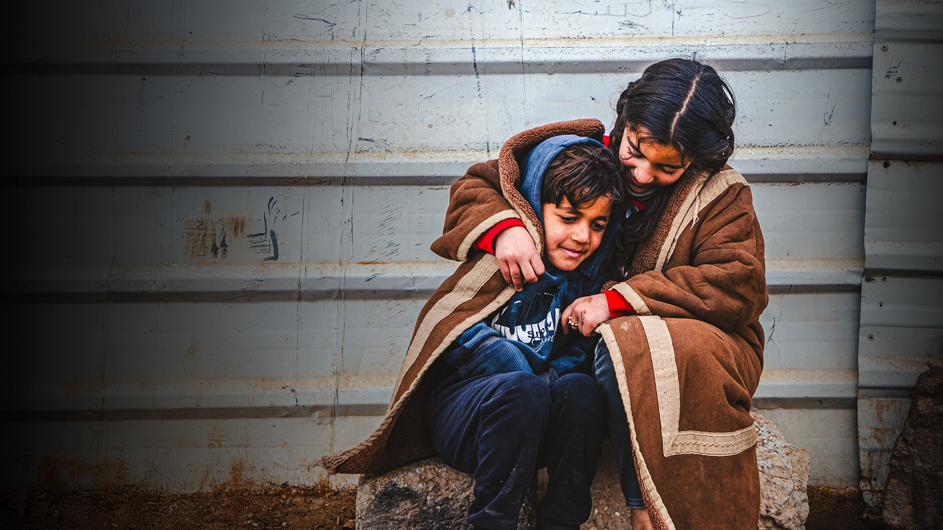 A young Syrian refugee wraps herself and her younger brother in a blanket as they sit outside in the cold in the Zaatari refugee camp.