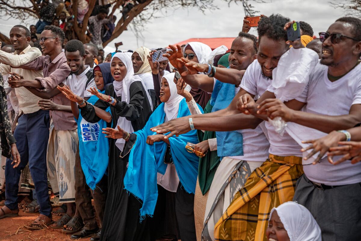 A large group of people dance outdoors while others others sit in a tree nearby