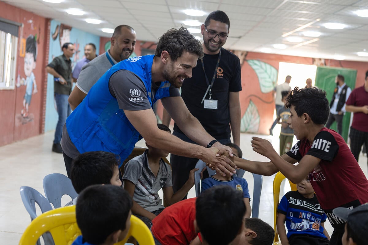 A man wearing a blue vest shakes hands with one of a group of children inside an