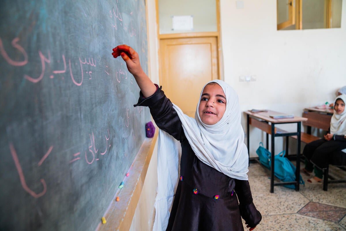 An Afghan girl holds up her arm towards a blackboard in a classroom, as others sit at desks behind her.