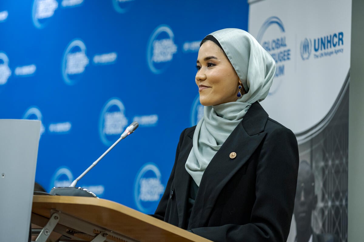 A woman speaks into a microphone at a podium in front of a UNHCR/Global Refugee Forum banner.