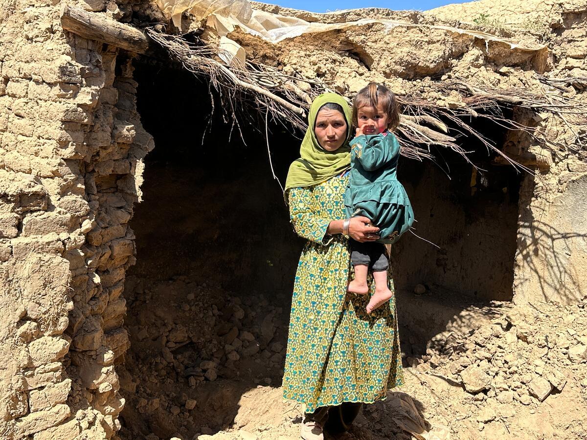 A woman carrying a young girl stands in front of a partially collapsed mud-brick building