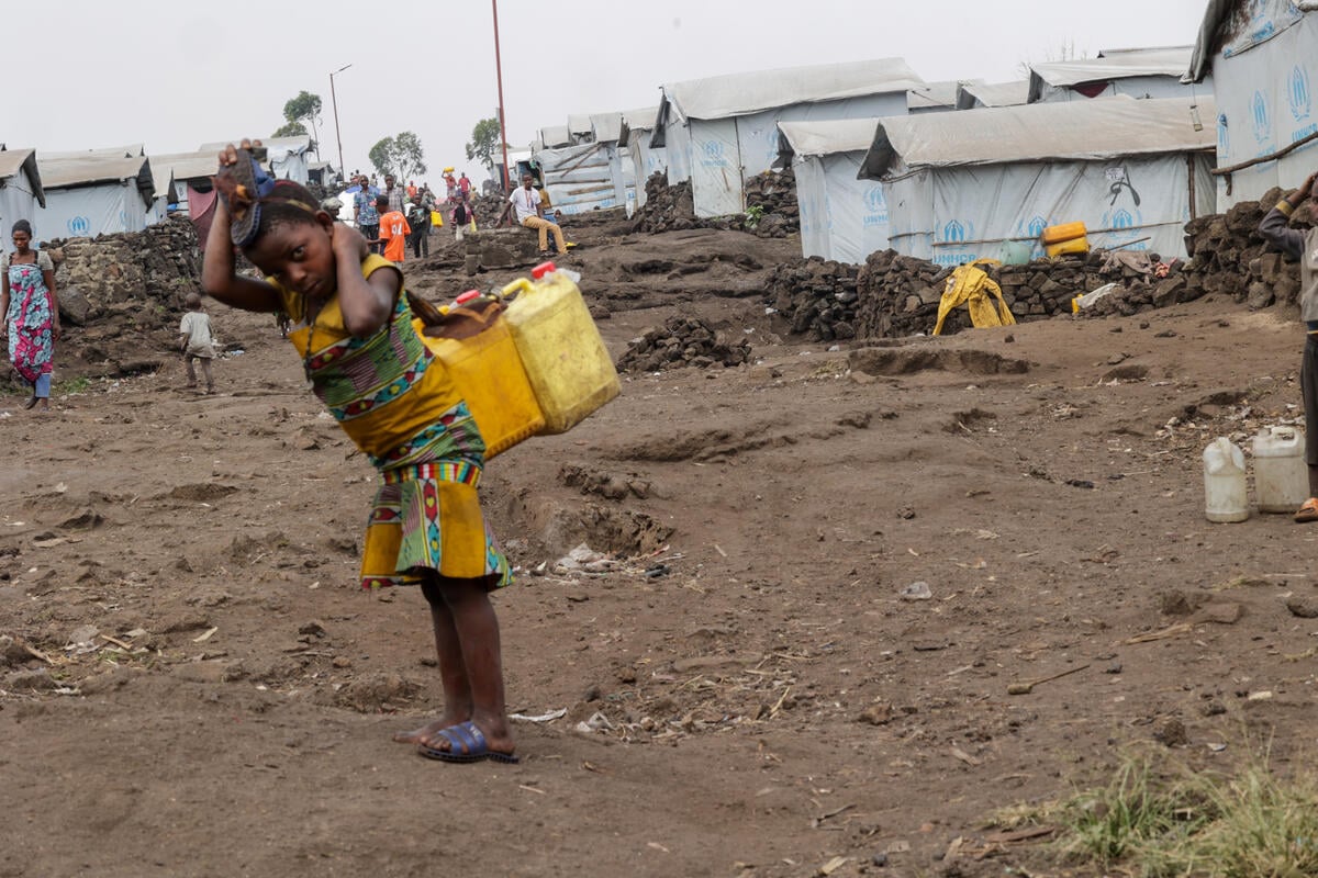 A young girl with two jerry cans on her back stands in front of a row of tents covered in tarpaulin.