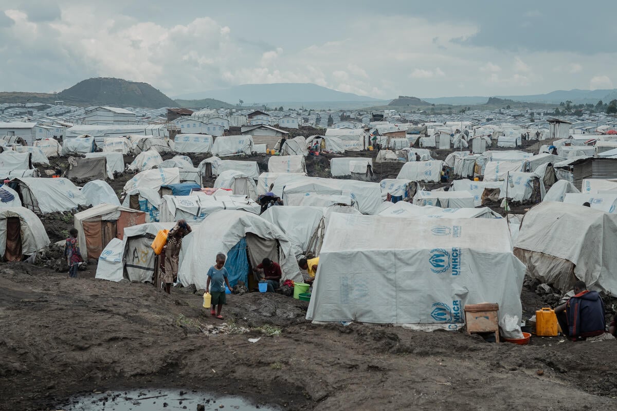 A young child walks across muddy ground in front of UNHCR tents with mountains in the background.