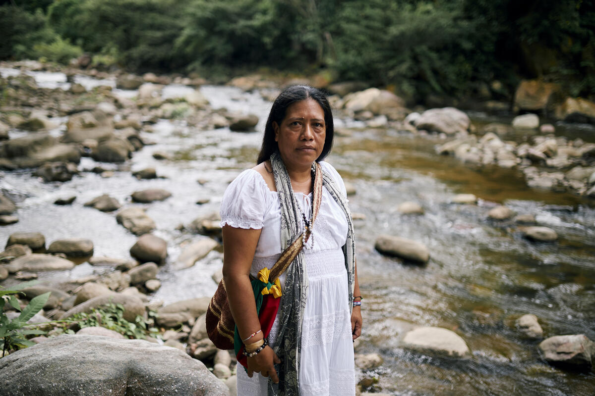 A woman wearing a long white dress with a colourful woven bag on her shoulder stands on the bank of a shallow rocky river