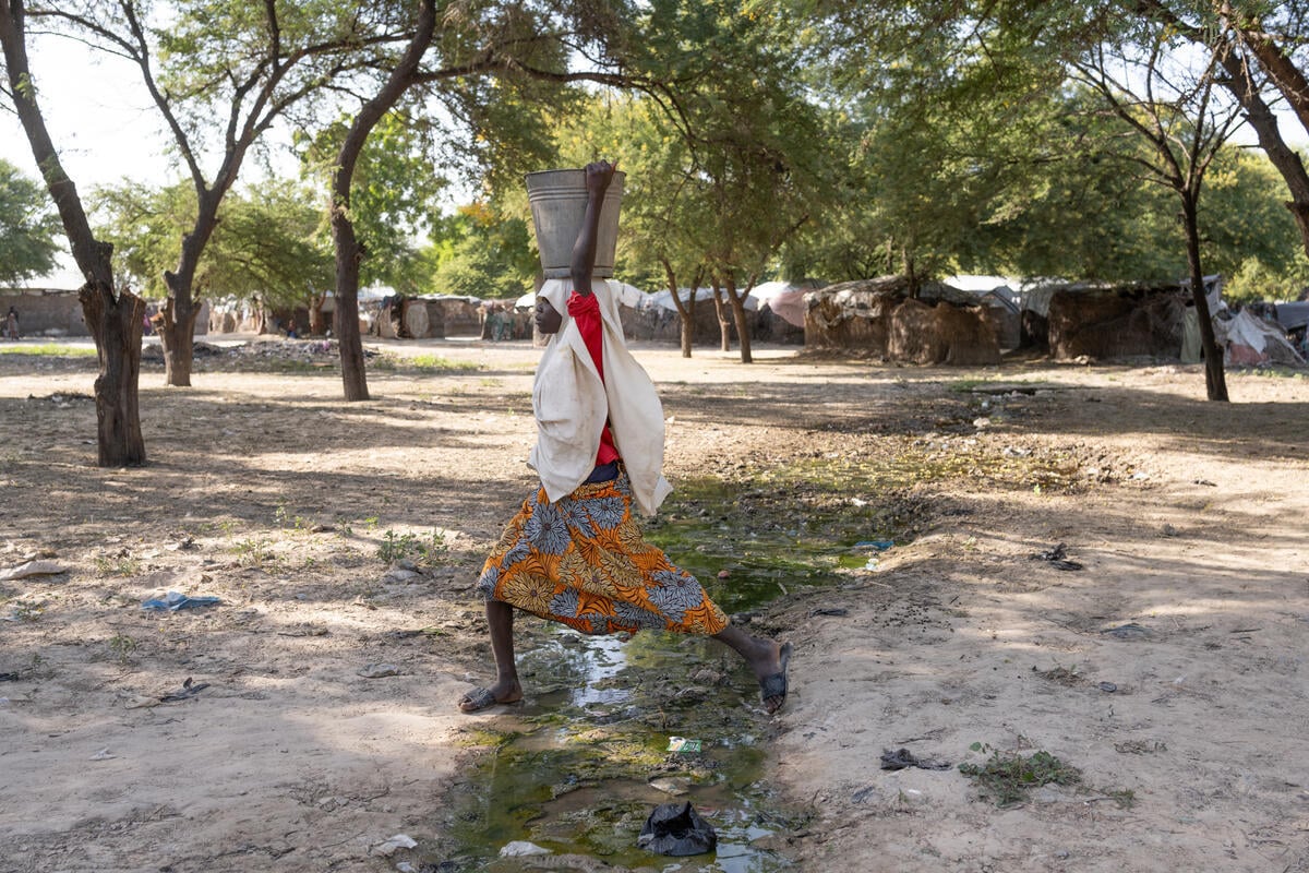 A girl holding a bucket on her head steps over a stream running through a camp.