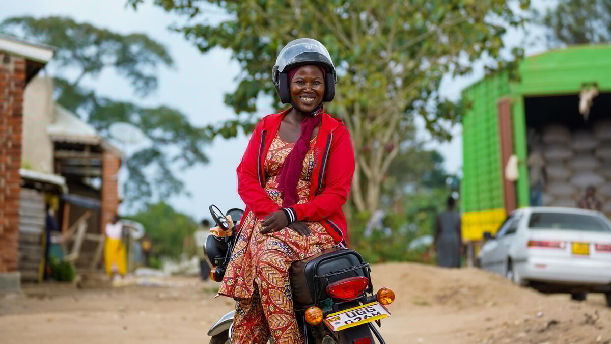 A woman sits sideways on a motorbike saddle wearing a helmet and smiling.