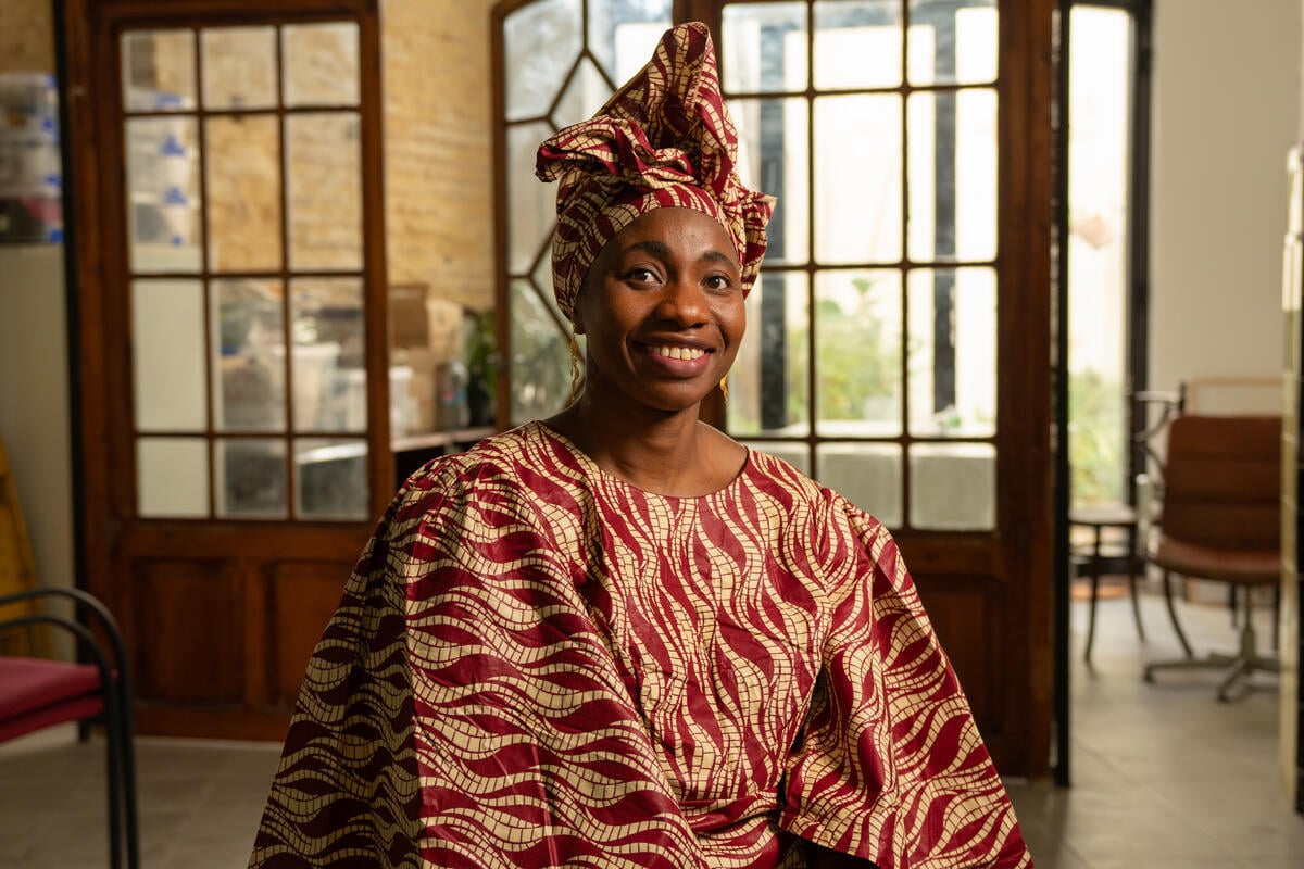 A smiling woman in a patterned traditional dress and headscarft sits in an office