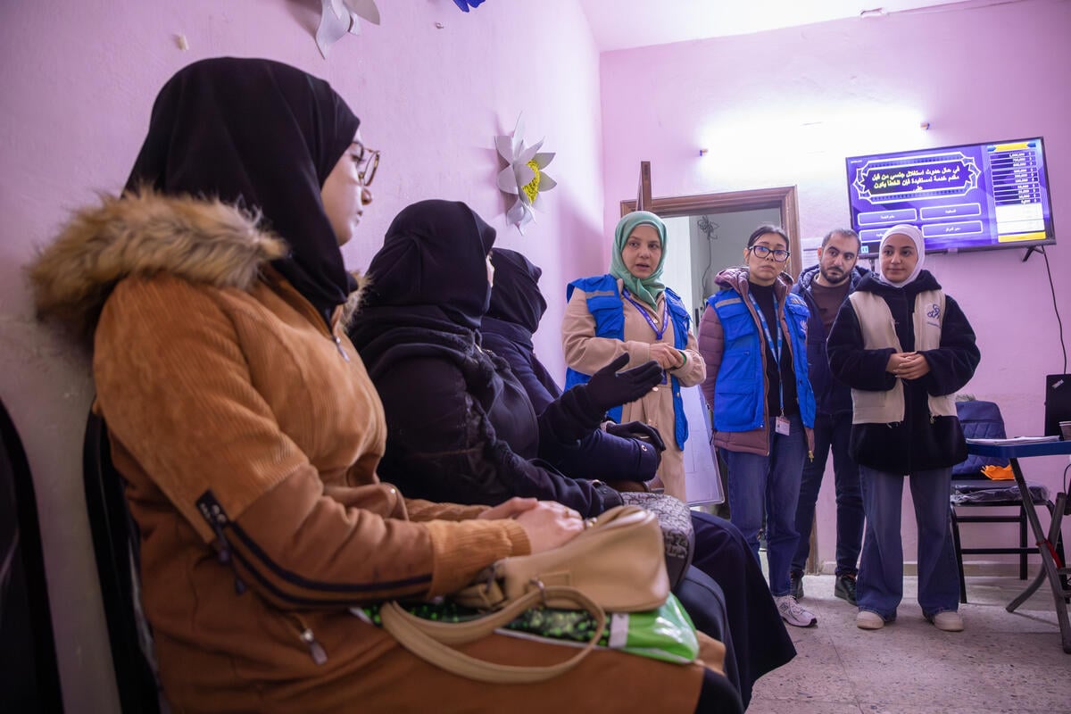 Women sit in a row at a community centre while UNHCR staff stand nearby, speaking to them