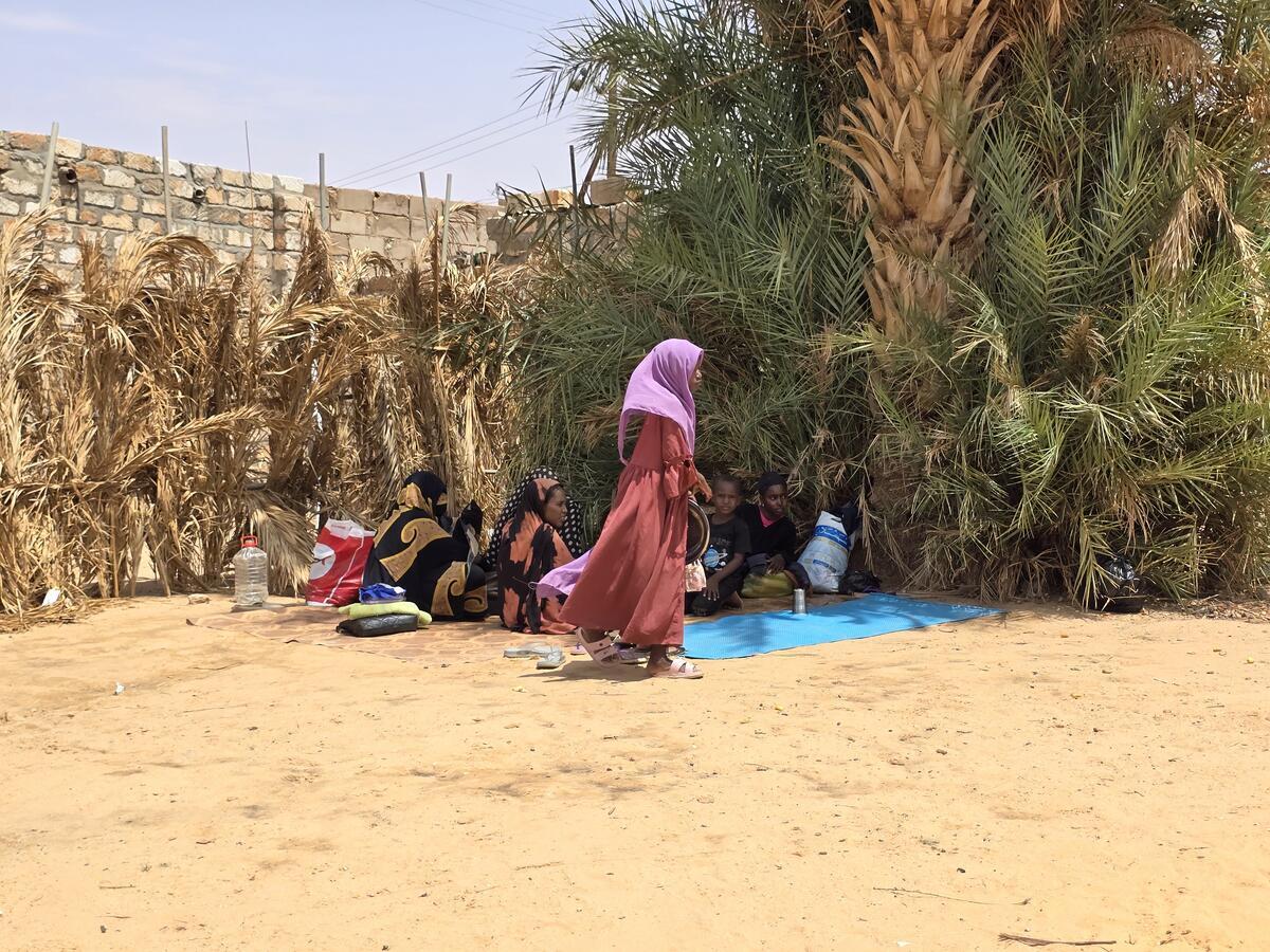 A Sudanese woman walks by a group of people sitting in the shade of some large trees in a dry environment.
