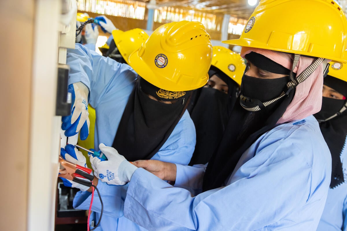 Two women wearing niqabs and yellow hardhats work together to screw a switch to a wall while other women look on.