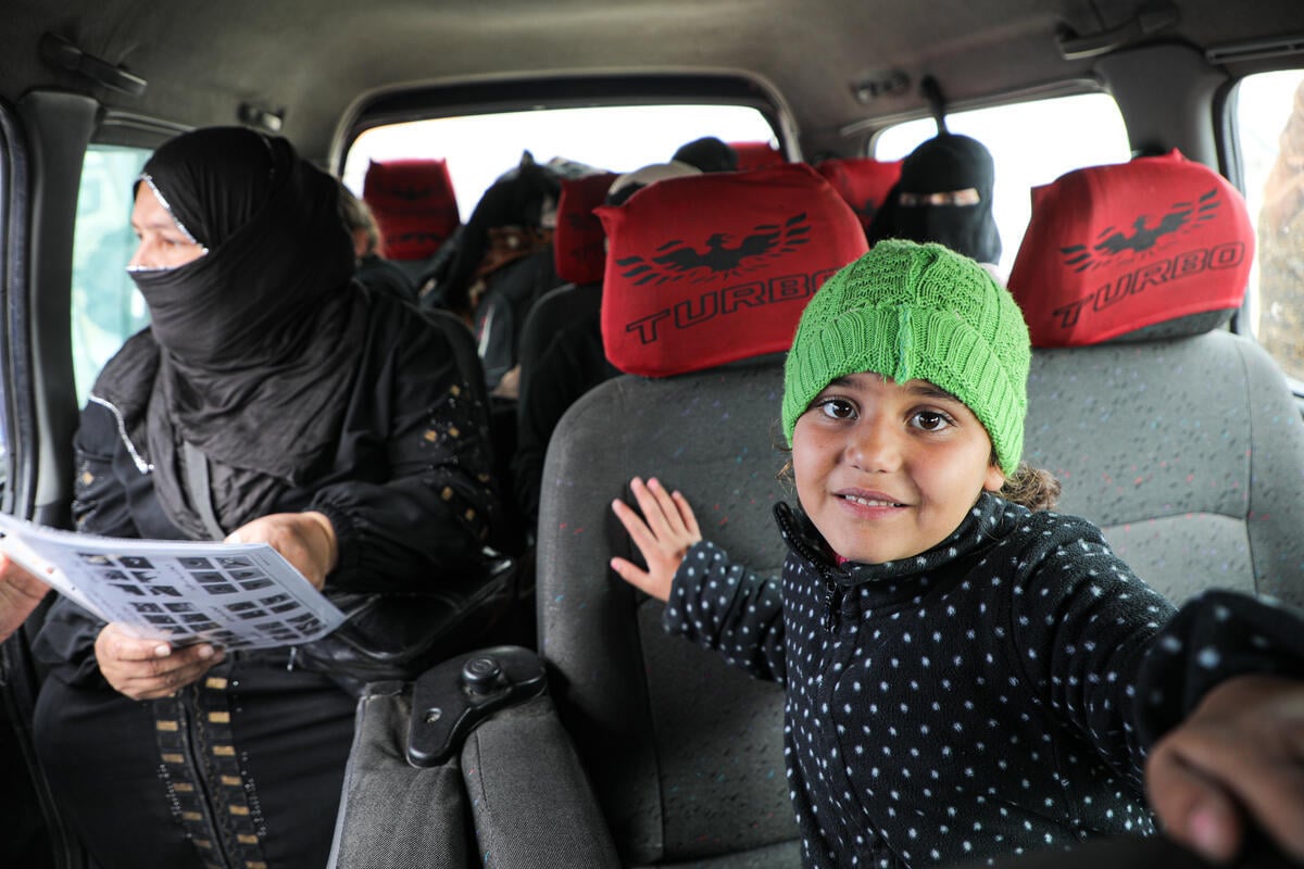 A young girl wearing a knitted green hat sits inside a minibus with women passengers sitting in the seats behind her.