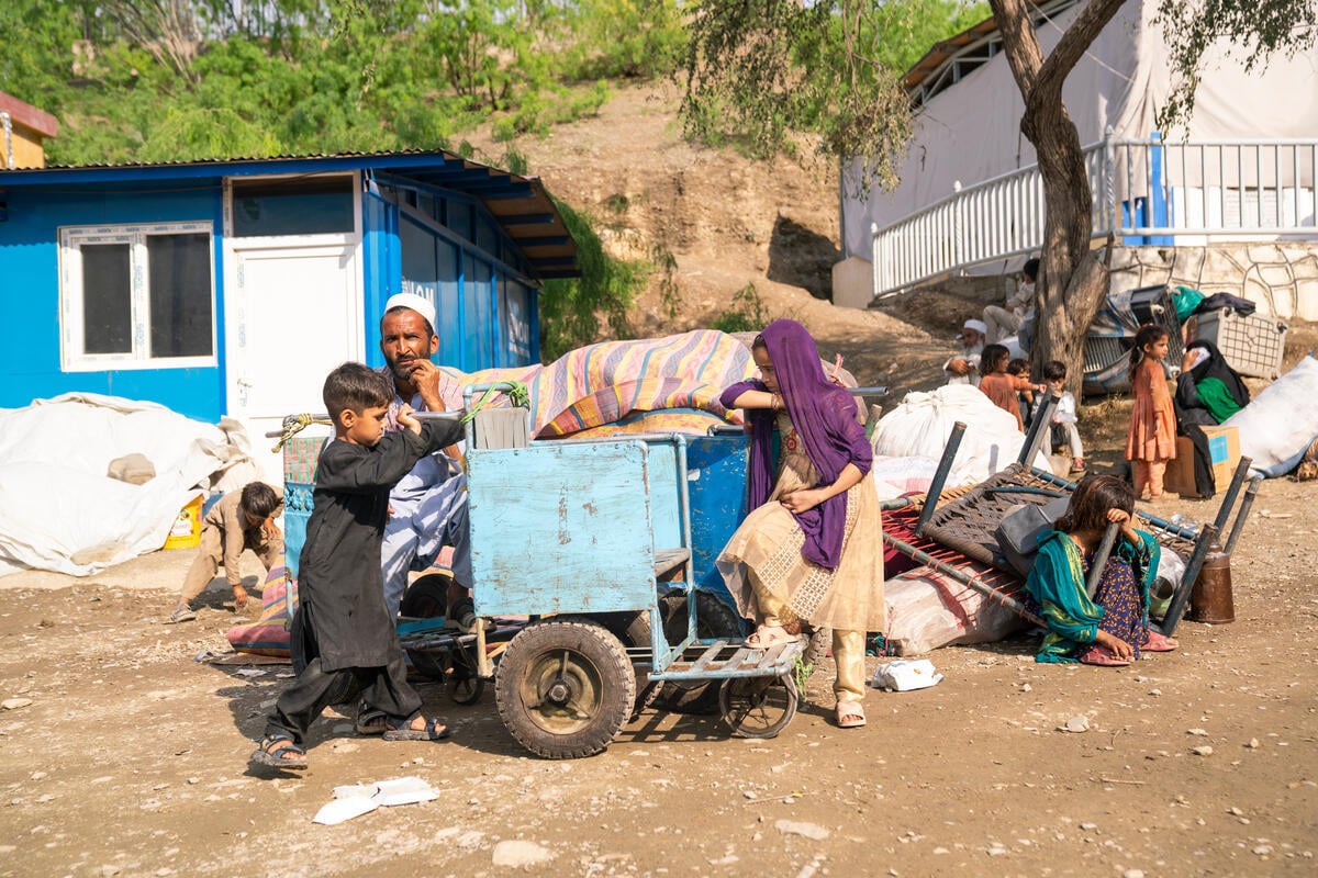A man and several children sit and stand near their belongings outside in front of a shelter.