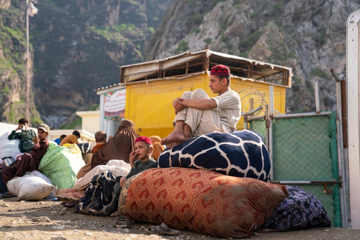 Two brothers wait at a bus stop in front of the mountains, sitting on large piles of belongings.