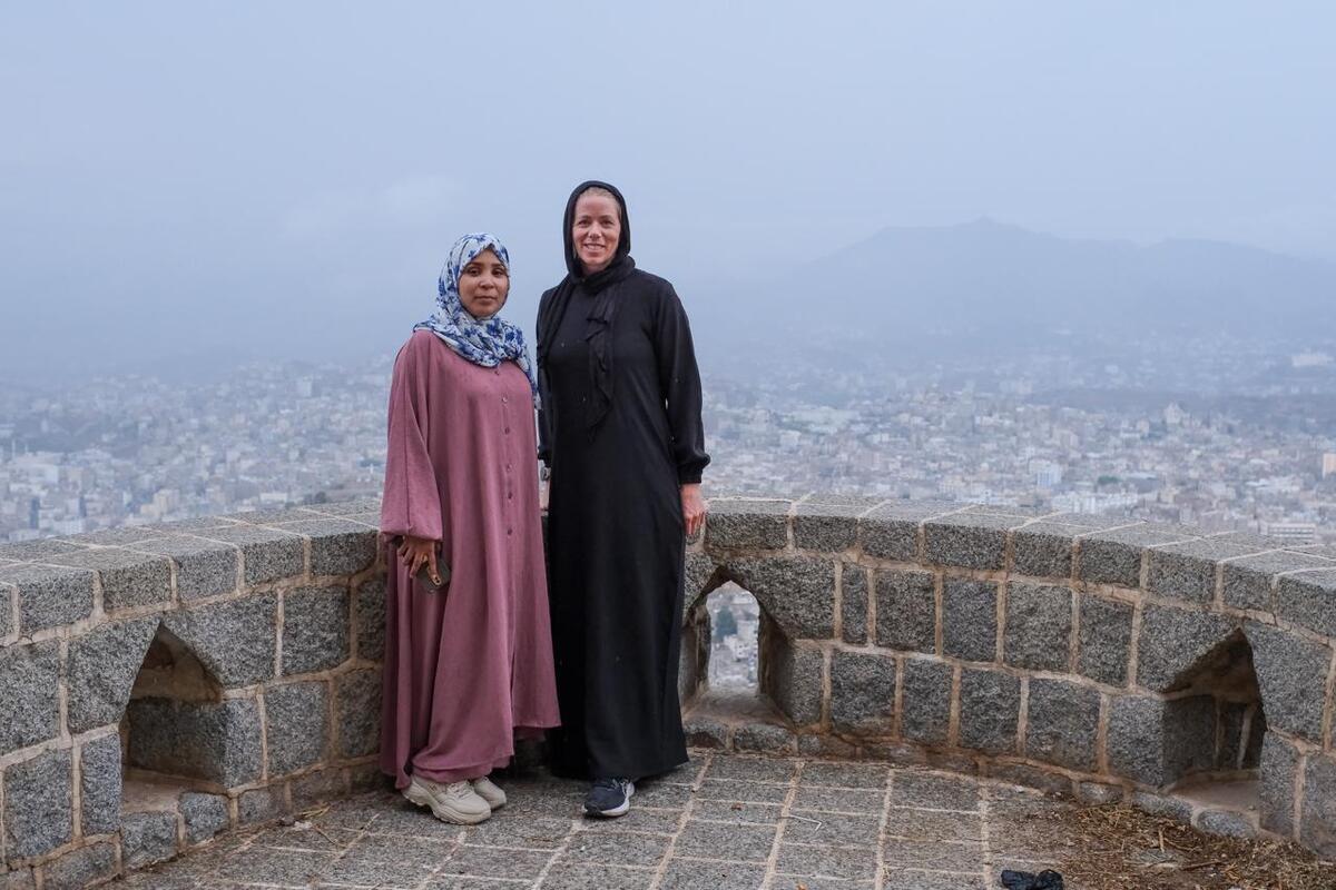Two women stand on a stone terrace with a city landscape behind them