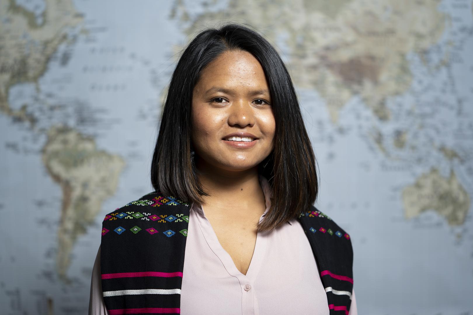 woman with shoulder-length dark hair, pictured from the shoulders up, smiling and standing in front of world map