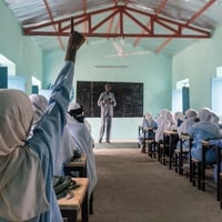 A rear view of a pupil raising her hand at the back of a classroom filled with schoolgirls in uniform sitting at rows of desks 