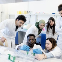 A group of people wearing lab coats gather round as a student and professor carry out an experiement in a laboratory 
