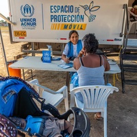A female member of UNHCR staff speaks to a woman across a table with a baby stroller in the foreground and a truck in the background