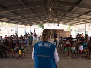 A woman in a branded UNHCR vest addresses a group of people in an open-sided building.