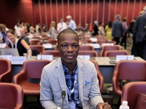 A man sits in a conference room with other delegates in the background.