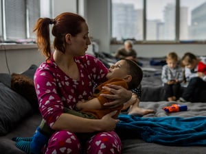 A woman sits on a camp bed holding her young son in a collective shelter for refugees.