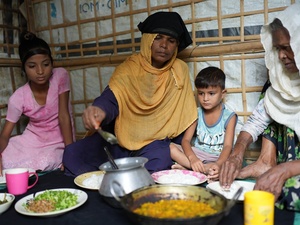 A mother and a grandmother eat a meal sitting on the floor of their shelter with two young children.