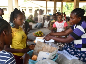 At a market, a woman hands over a small baguette wrapped in paper to a customer.