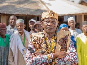 A man in colourful robes with his arms crossed stands in front of a group of people