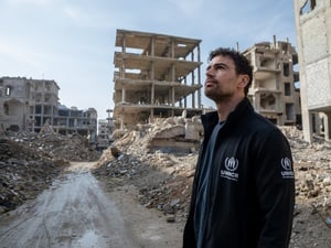 A man wearing a black UNHCR jacket looks upwards while surrounded by rubble and destroyed buidings