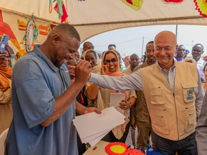 A smiling man drawing a pencil portrait on paper receives a pat on the shoulder from his smiling subject in a tent full of onlookers