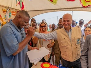A smiling man drawing a pencil portrait on paper receives a pat on the shoulder from his smiling subject in a tent full of onlookers