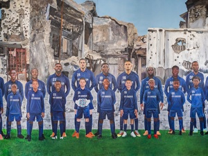 A painting showing a line-up of football players with their younger selves standing in front of them, with war-damaged buildings in the background