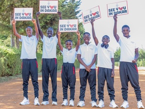 Six smiling young athletes wearing training shoes and clothes hold up signs reading 'see you in Dakar' on a wide dusty road 