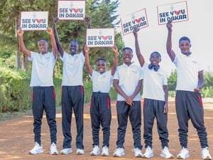 Six smiling young athletes wearing training shoes and clothes hold up signs reading 'see you in Dakar' on a wide dusty road 