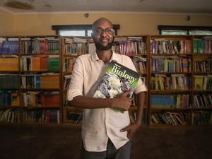 Abdullahi Mire stands in a library in front of a huge bookshelf, clutching a biology book