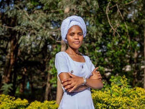 A woman in a blue dress and headscarf stands outside with her arms crossed and trees in the background