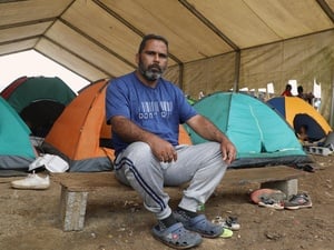 A man sits on a low bench in front of some tents.