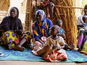 Women and children sit on a mat on the floor in a shelter.