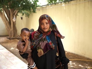A woman wearing a headscarf stands in a walled courtyard holding a toddler.