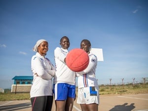 Three women stand together in a basketball court, one holding out a basketball.