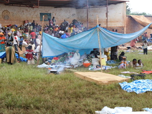 Large groups of people gather with their belongings under a large outdoor structure and smaller makeshift shelters