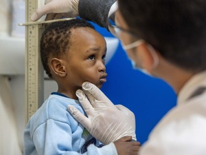 A young boy in a blue sweater stands against a height rule as a man wearing a surgical mask and gloves measures his height.