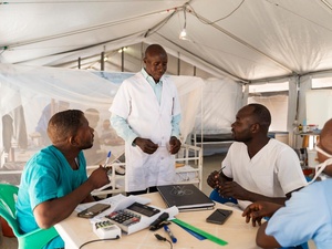 A male nurse in a white coat talks to his colleagues who sit at a round table in a hospital