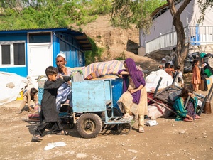 A man and several children sit and stand near their belongings outside in front of a shelter.