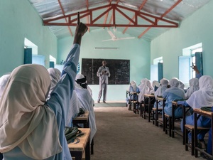 A rear view of a pupil raising her hand at the back of a classroom filled with schoolgirls in uniform sitting at rows of desks 