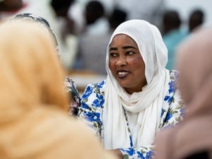 A smiling woman in a white headscarf and blue floral dress sits at a table with other women