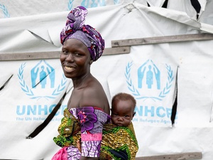 A smiling woman carries her baby boy on her back in front of a UNHCR tent.